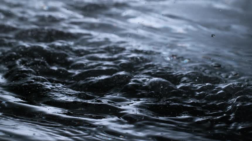A macro slow-motion shot captures the mesmerizing movement of dark, bubbling liquid as a single droplet forms and falls back to the surface