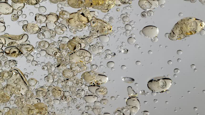 A macro close-up of golden oil bubbles slowly floating and clustering together in clear water against a white background