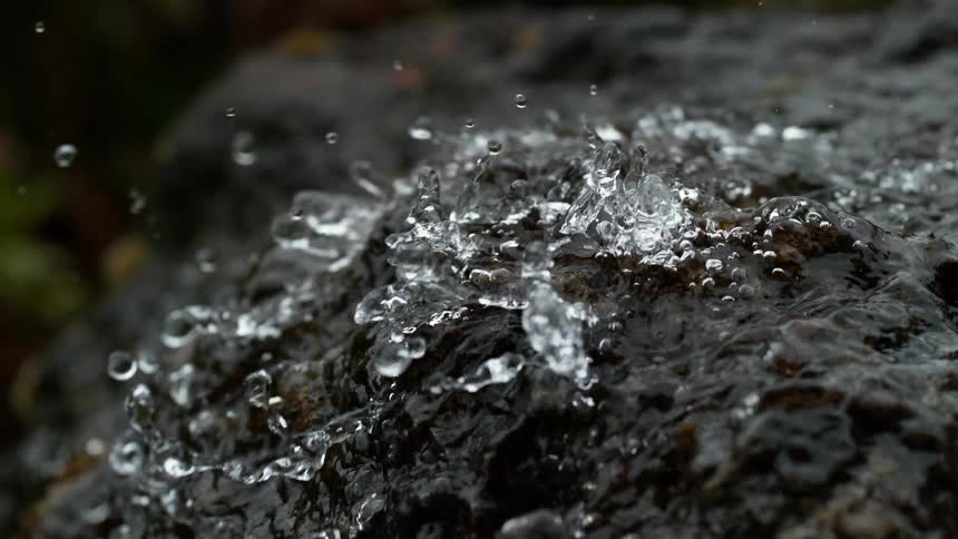 Super slow motion close up of clear water splashing and bubbling on a dark wet rock