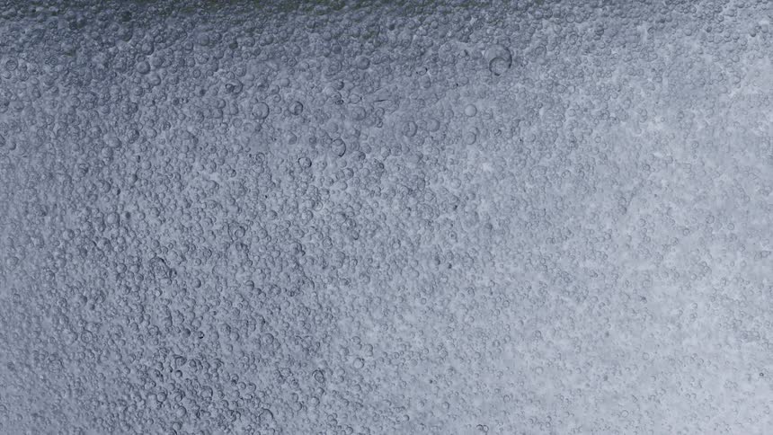 A macro close-up of a viscous liquid with tiny air bubbles slowly rising to the surface against a grey background