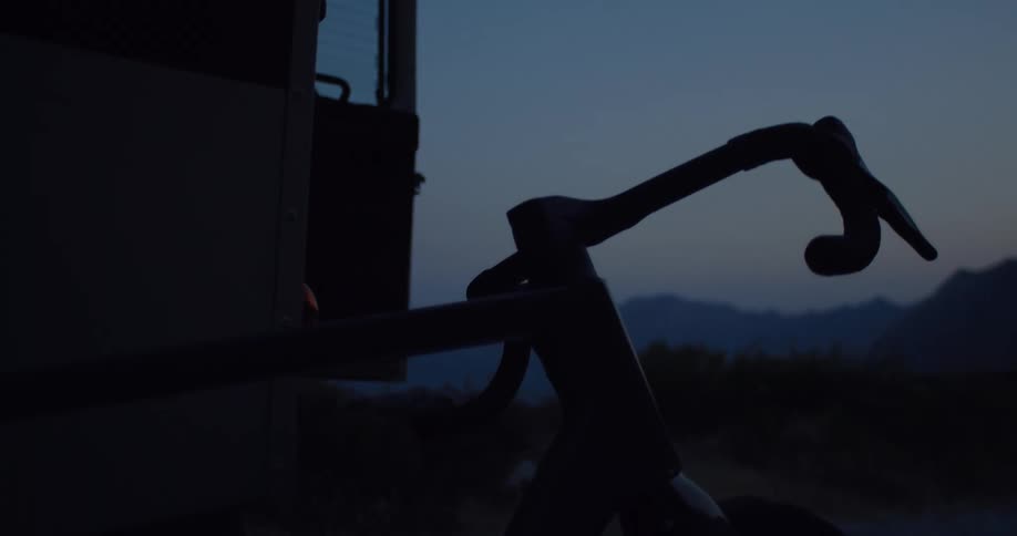 A close-up shot of a silhouetted bicycle resting next to a van at dusk with mountains in the background