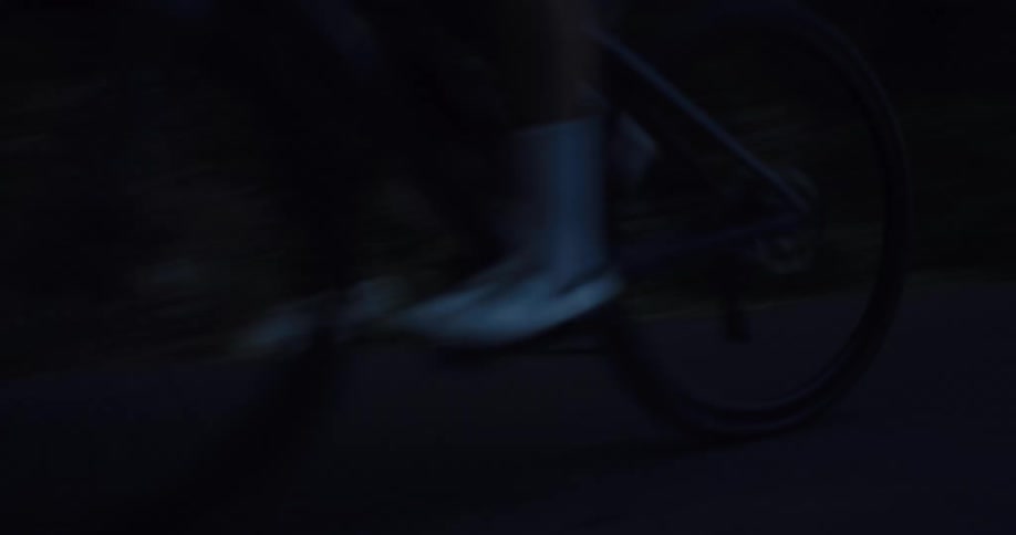 Low-angle shot of a silhouetted cyclist riding a road bike along a dark, tree-lined road at dusk