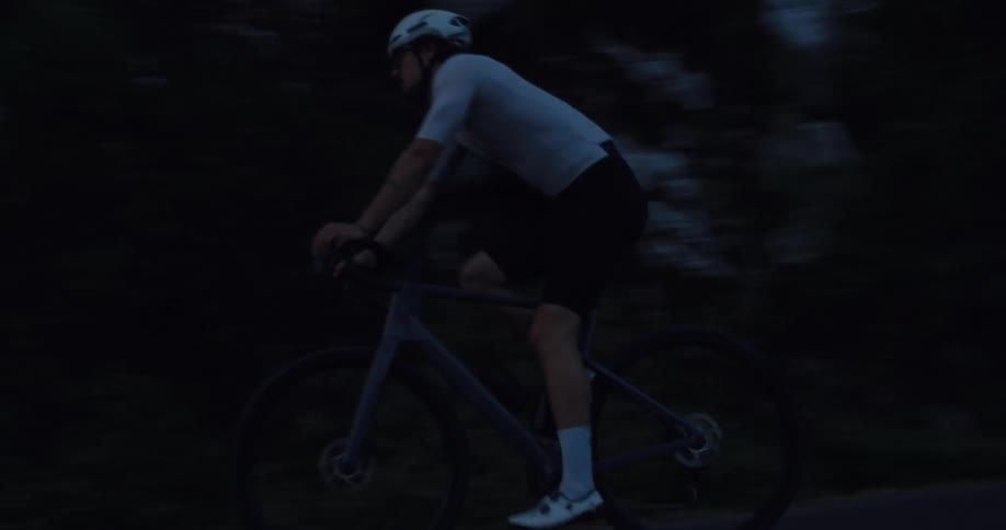 A male cyclist in a helmet rides his bike on a dark, winding road at dusk