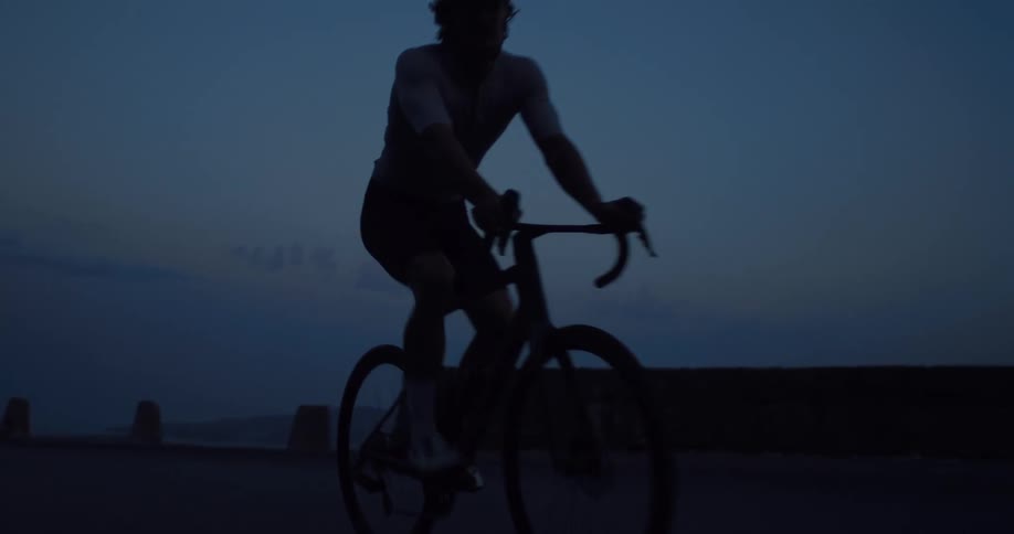 A silhouetted cyclist rides his road bike along a mountain pass at dusk in slow motion