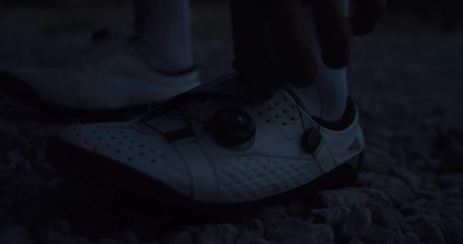 A cyclist fastens their specialized white cycling shoes on a gravel path in the dark before a ride