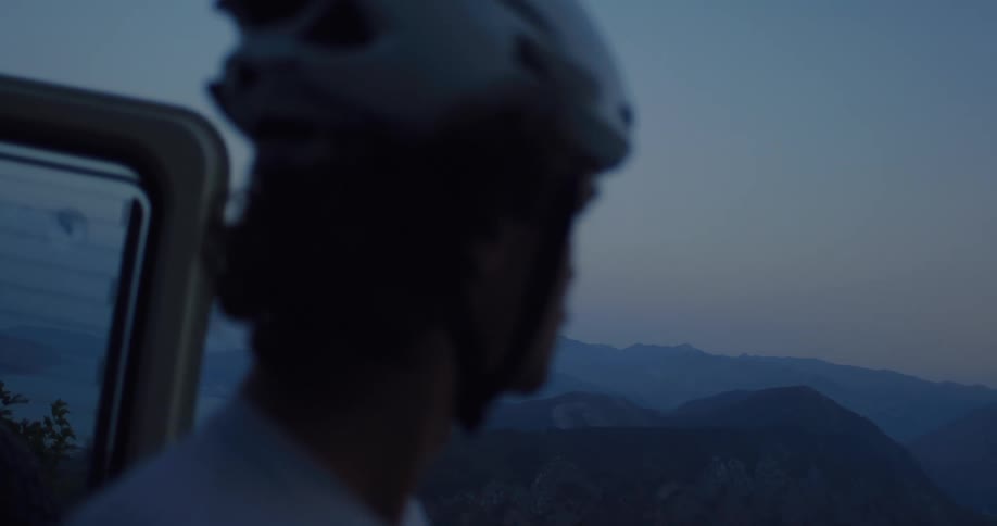A silhouetted person wearing a helmet looks out at a vast mountain range at dusk