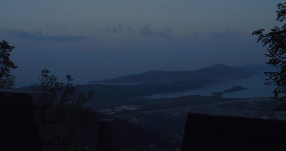 A cyclist is silhouetted as they ride past a scenic mountain viewpoint overlooking a bay and coastal city at dusk