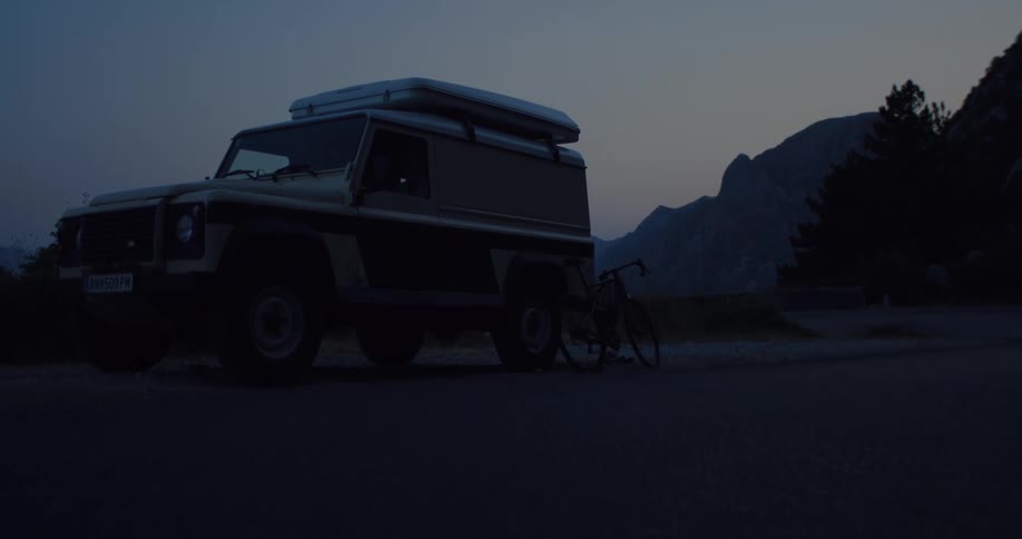 A slow push-in shot captures an off-road vehicle with a rooftop tent and bicycle parked on a mountain road at twilight