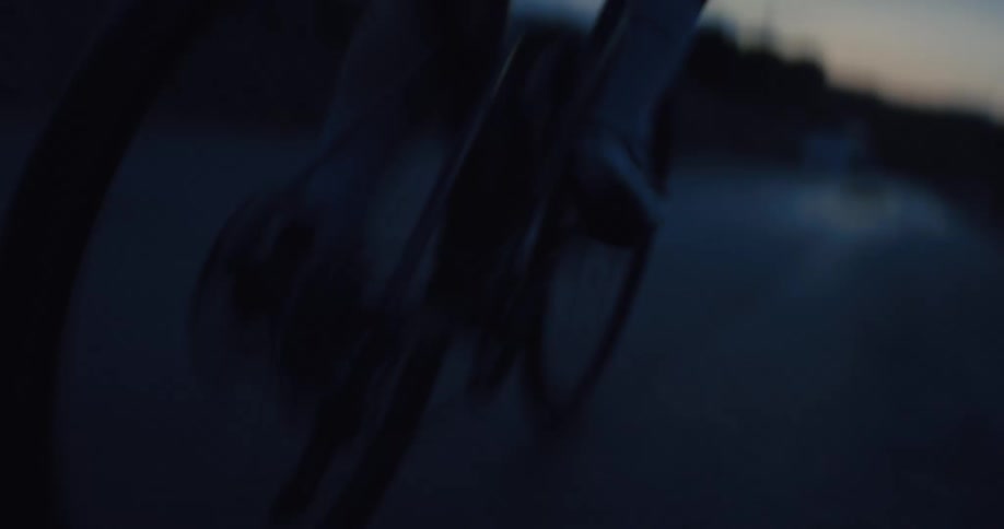A close-up, out-of-focus shot follows a cyclist's legs and spinning wheels as they ride on a road during the blue hour at twilight