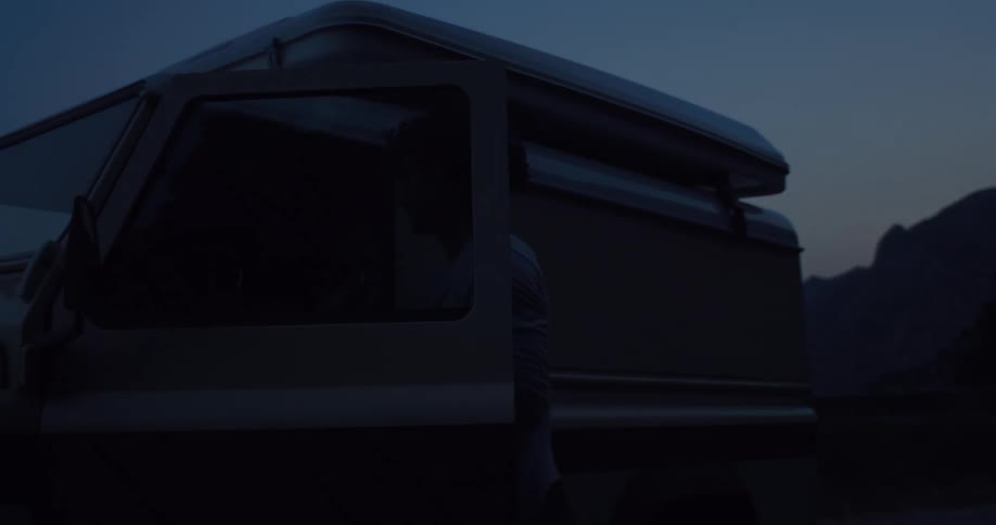 A silhouetted man gets out of his vintage camper van at dusk in a remote mountain landscape in slow motion