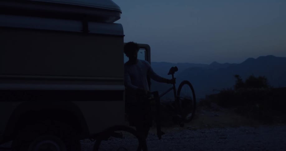 A silhouetted man slowly unloads his bicycle from his off-road camper van at dusk with a beautiful mountain view