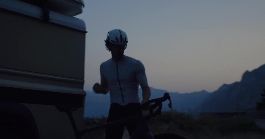 A professional cyclist in silhouette prepares his bike for a ride next to a camper van in the mountains at twilight