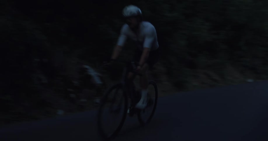 Tracking shot of a male cyclist riding his bike along a dark, forested road at twilight
