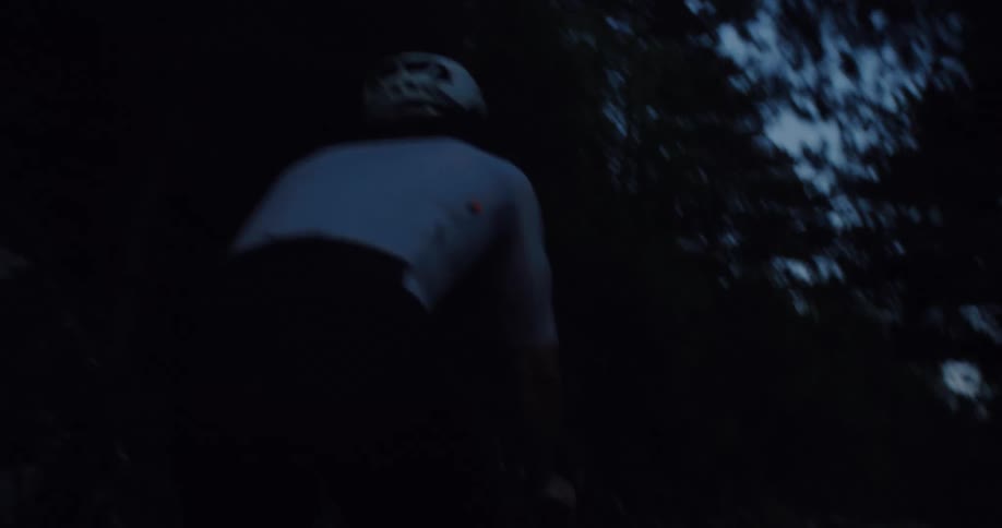 A low-angle, shaky shot follows a cyclist as they pedal hard uphill through a dark forest at twilight