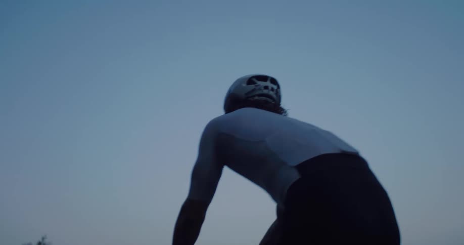 A low-angle, follow-shot of a professional road cyclist pedaling hard up a mountain road during the blue hour