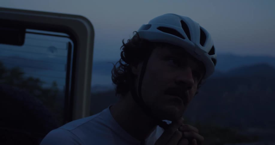 Slow motion close-up of a determined male cyclist putting on and adjusting his helmet at dusk, with mountains in the background