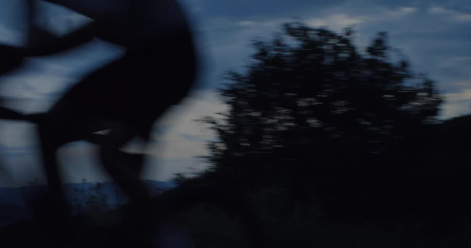 A cyclist rides swiftly past the camera on a dark country road at dusk, revealing a scenic mountain landscape