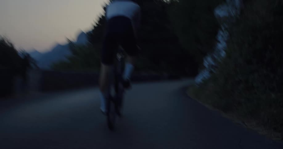 A professional cyclist in athletic gear rides a road bike along a secluded mountain road at dusk