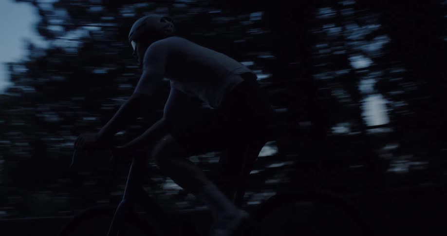 A professional cyclist rides a dark, winding mountain road in a moody slow-motion tracking shot at dusk