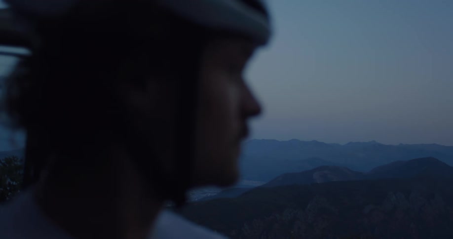 A contemplative cyclist in a helmet looks out over a vast mountain range during the tranquil twilight hours