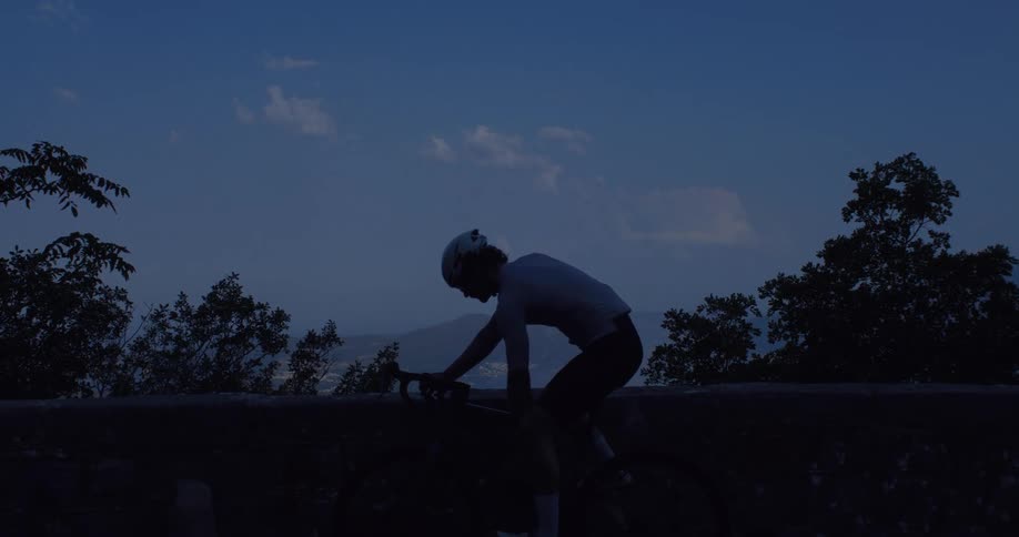 A silhouetted cyclist takes a break for a drink at dusk, overlooking a scenic mountain and water landscape before riding away