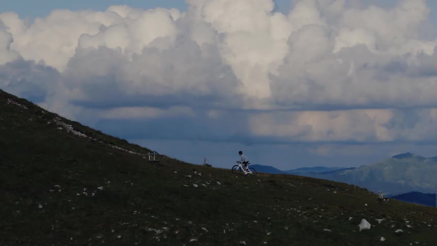 An aerial shot slowly approaches a lone mountain biker pushing their bike up a vast, grassy slope under a dramatic cloudy sky