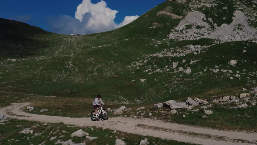 An aerial view of a person riding a gravel bike along a dirt trail in a vast, green mountain landscape under a blue sky