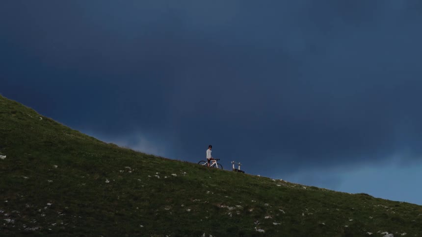 A lone cyclist stands on a green mountain pass, contemplating the view under dark and stormy skies
