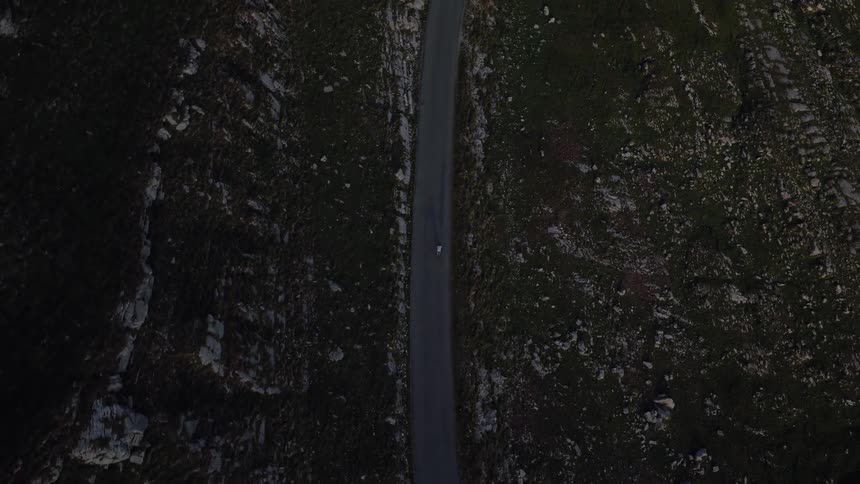 A lone white car travels down a remote paved road surrounded by a vast and rugged rocky landscape in a dramatic top-down aerial view