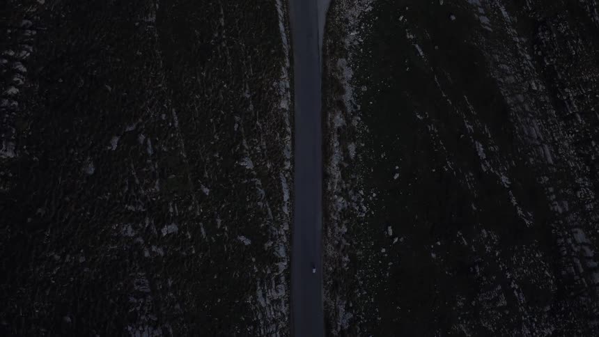 A moody top-down aerial view of a lone white car driving on a remote road through a dark and desolate mountain landscape