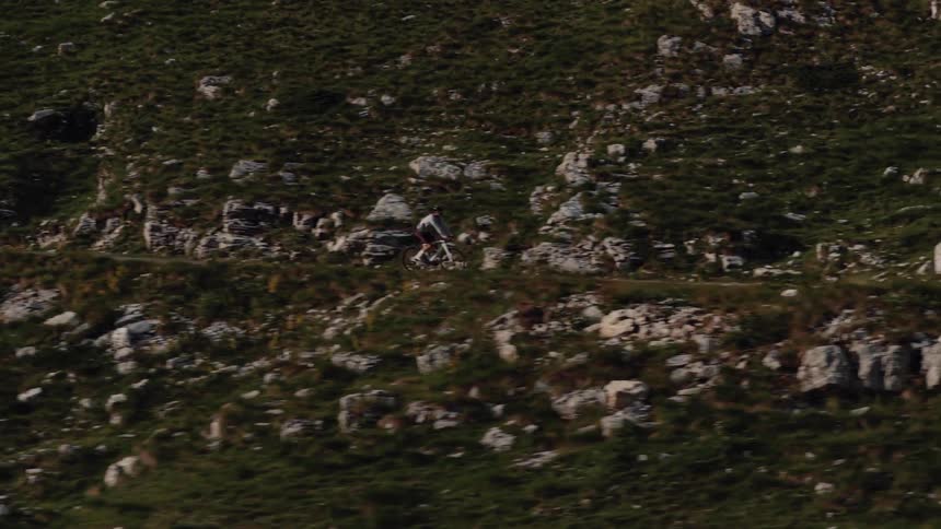 An aerial tracking shot follows a lone cyclist riding along a narrow path on a steep, rocky, and green mountainside