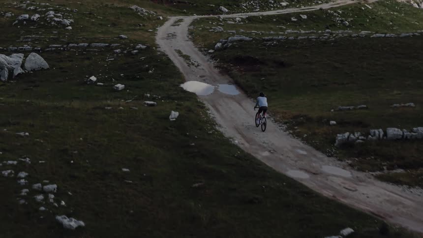 An aerial slow-motion shot of a lone cyclist riding a mountain bike along a remote dirt trail in a vast, rocky, and green mountain landscape