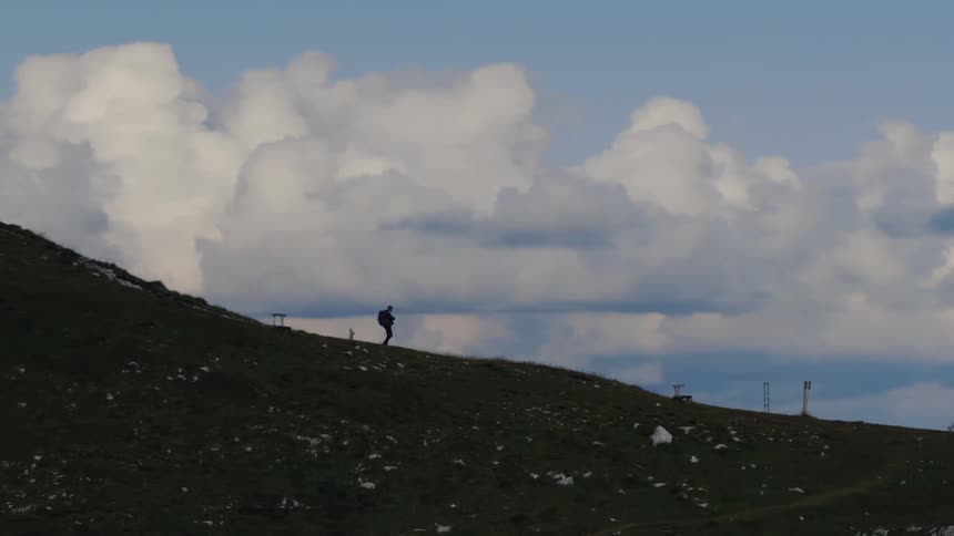 A silhouetted hiker with a backpack and poles makes their way up a grassy mountain ridge against a backdrop of large, fluffy clouds