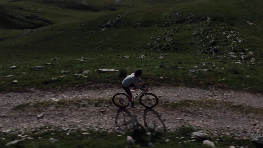An aerial side view tracks a male cyclist pedaling uphill on a gravel trail through a vast, green mountain landscape