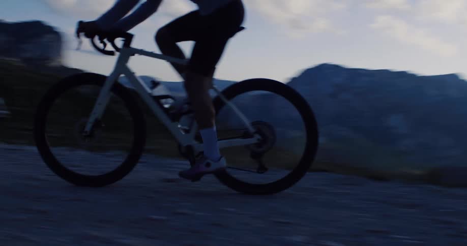 A cyclist rides up a winding gravel mountain road at dusk, silhouetted against a dramatic cloudy sky