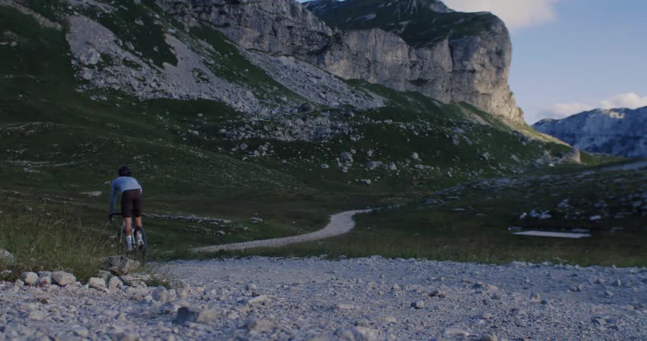 A cyclist rides down a winding gravel path into a beautiful mountain valley at golden hour