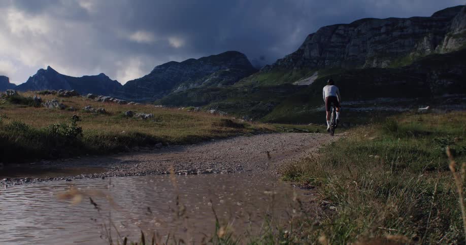 A cyclist rides through a large muddy puddle on a remote gravel mountain trail in cinematic slow motion