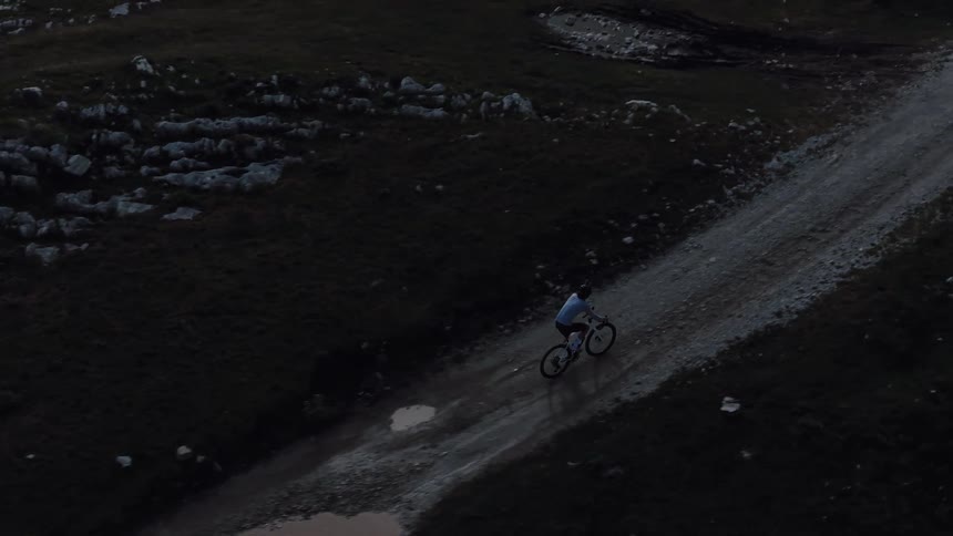 A lone mountain biker slowly ascends a steep gravel road in the dim light of dusk, seen from an aerial perspective