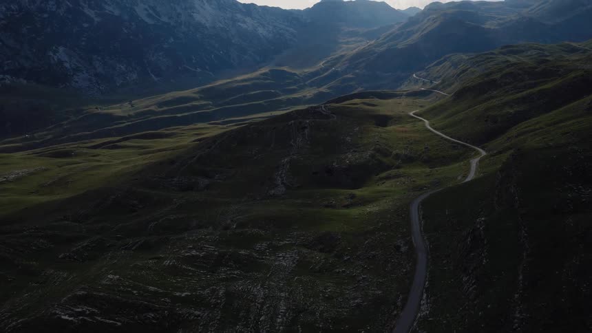 An expansive aerial shot follows a winding road through a lush green mountain pass as cars navigate the route towards towering peaks