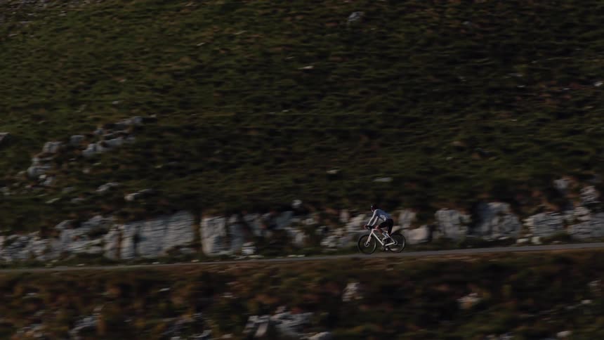 A lone cyclist pedals along a winding road carved into a steep, grassy mountainside in a smooth tracking shot