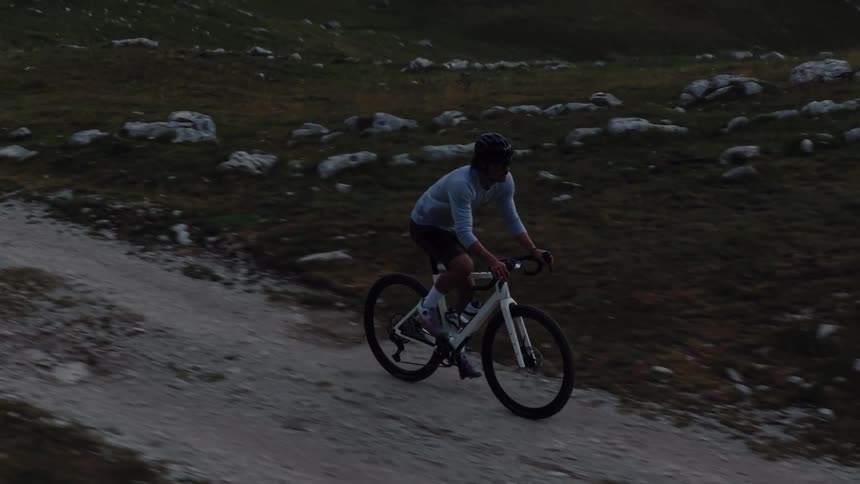 Aerial slow-motion tracking shot of a man riding a gravel bike uphill on a mountain path at dusk