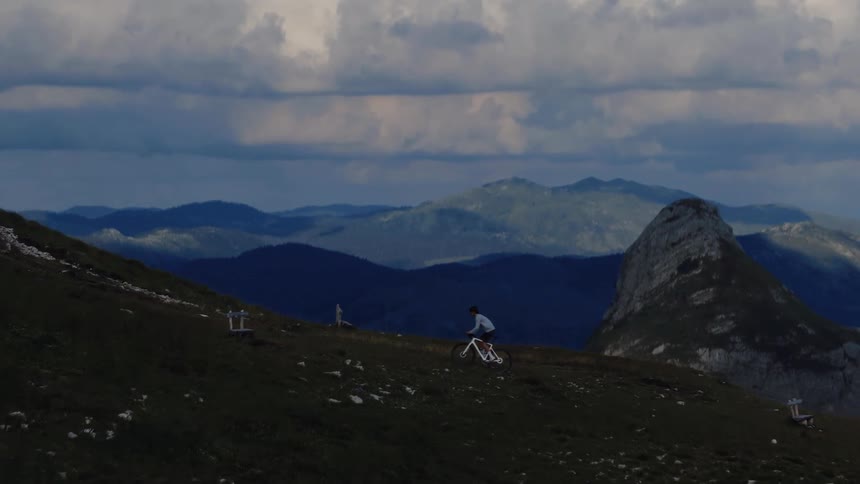 An aerial view follows a lone mountain biker traversing a grassy alpine slope against a backdrop of a vast mountain range and dramatic clouds