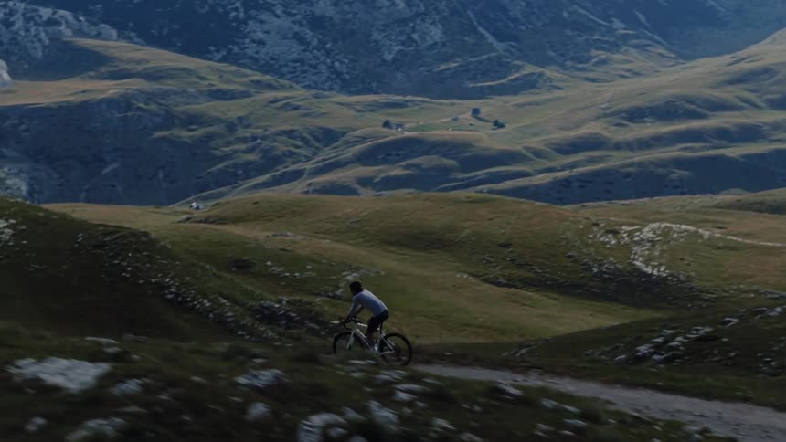 An aerial view follows a cyclist riding along a rugged dirt trail through a vast and scenic mountain landscape