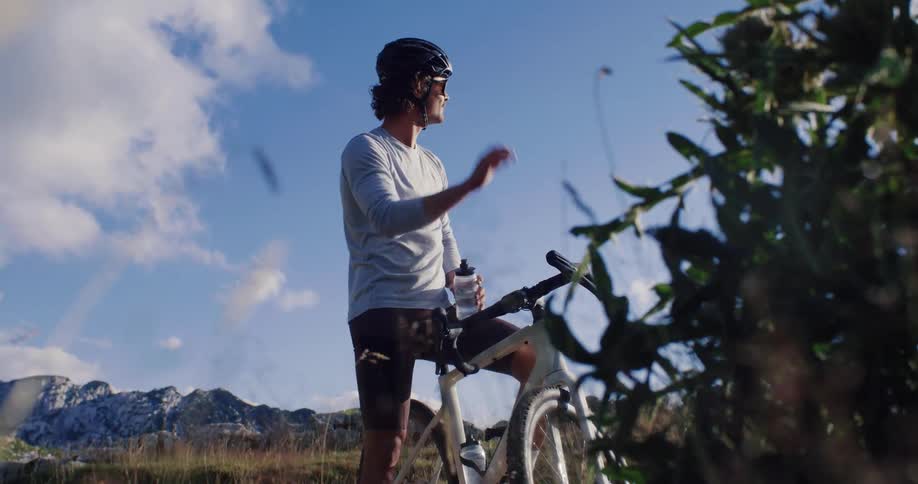 Low-angle view of a male cyclist taking a break to drink water while admiring the mountain landscape under a blue sky
