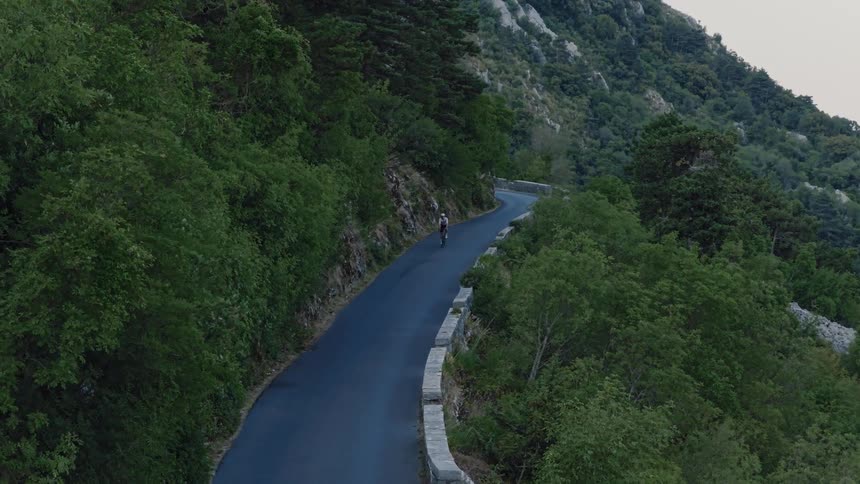 A lone cyclist rides up a winding mountain road surrounded by a dense, green forest