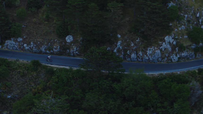 An aerial view follows a determined cyclist on a winding road carved into a steep, forested mountainside at dusk