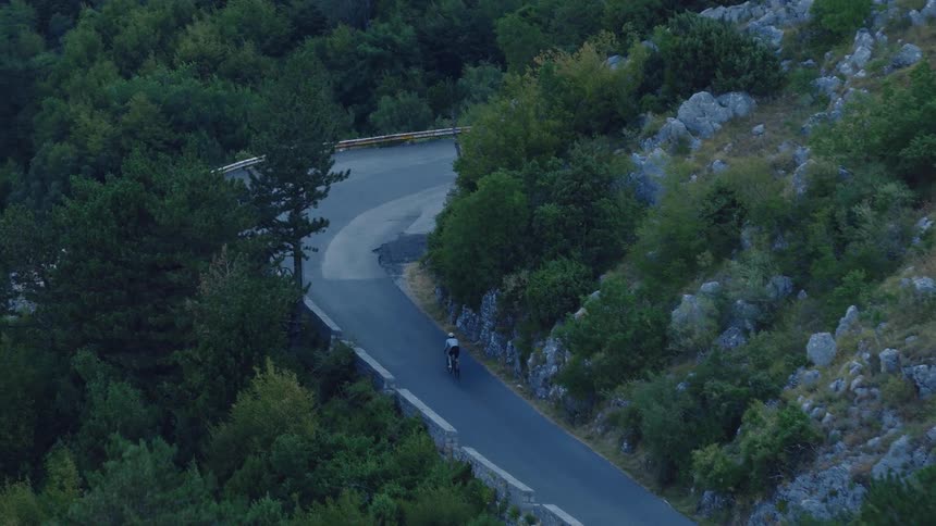 A lone cyclist pedals up a winding mountain road surrounded by a lush green forest and rocky terrain