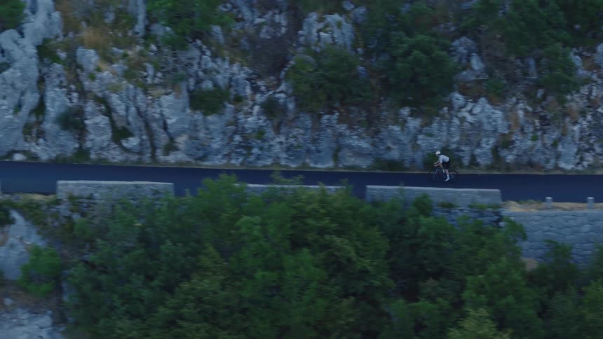 An aerial view captures a lone cyclist pedaling along a winding road carved into a rugged, tree-covered mountainside