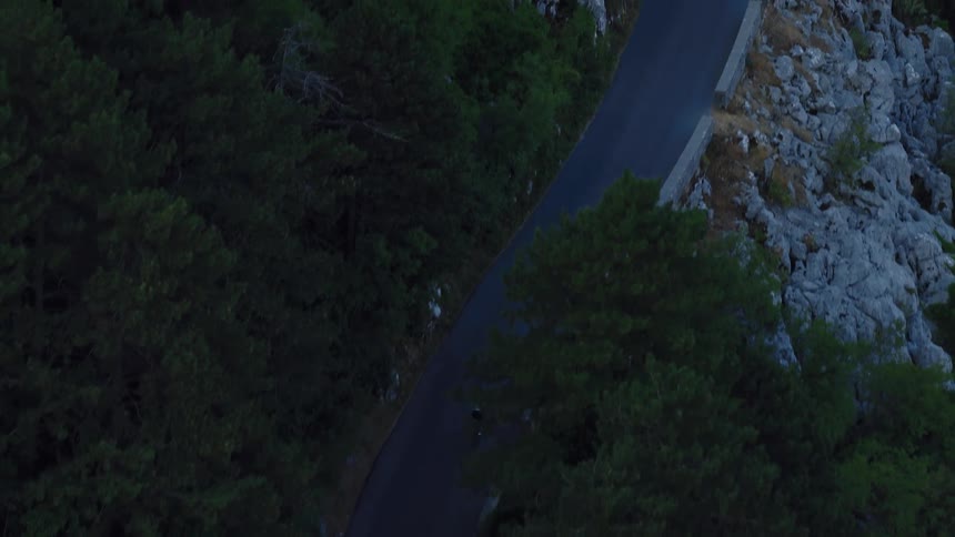 A high-angle aerial shot follows a lone cyclist riding up a winding road through a dark forest on a rocky mountainside
