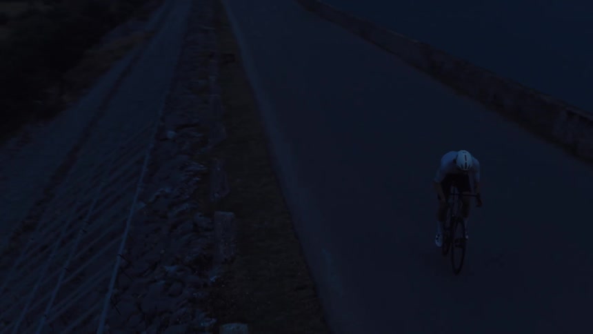 An aerial shot follows a lone cyclist riding on a dam road in the dark before dawn